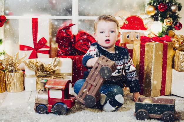 Retrato de niño rubio sentado en el suelo en estudio decorado y jugando con cajas y regalos de Navidad