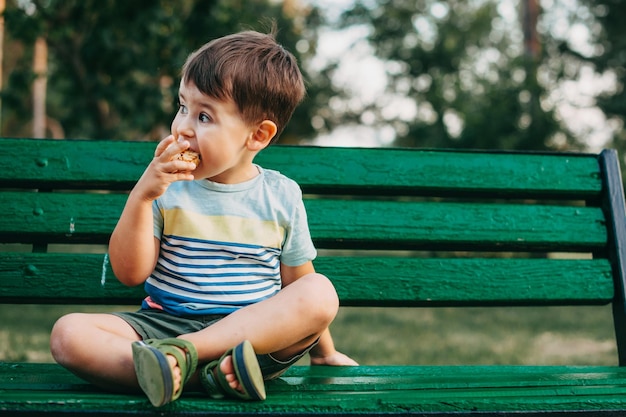 Retrato de niño sentado en un banco en el parque niño de pelo castaño años comiendo un pastel vista frontal