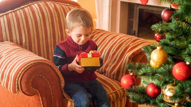 Retrato de niño sentado en un gran sillón y sosteniendo la caja de regalo de Navidad de Santa Claus