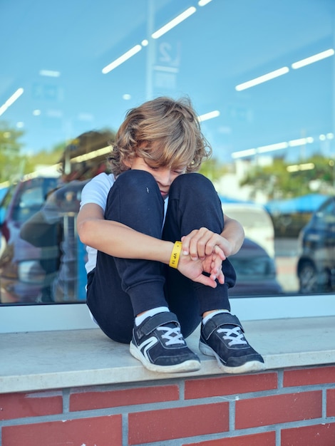 Retrato de un niño solitario sentado cerca de la ventana con autos estacionados reflejándose en ella y abrazando las rodillas mientras se siente molesto