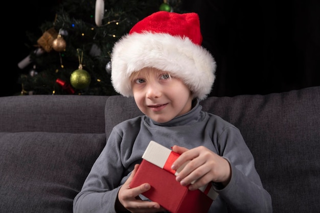 Retrato de niño con sombrero de Santa con regalo en sus manos. Niño sentado en el sofá con el telón de fondo del árbol de Navidad