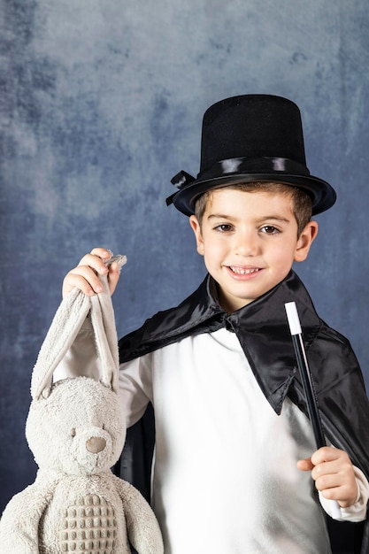 Retrato de un niño con sombrero sosteniendo un juguete de peluche y una varita contra un fondo gris