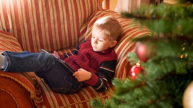 Retrato de niño sonriente acostado en el sofá junto al hermoso árbol de Navidad en la sala de estar y jugando en tableta digital