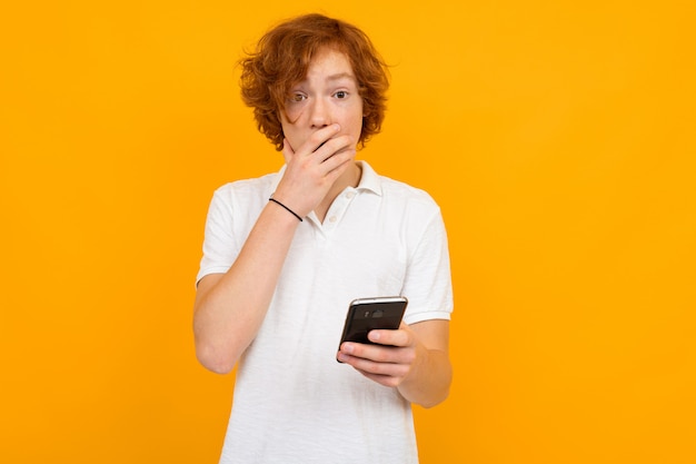 Retrato de un niño sonriente atractivo pelirrojo en una camiseta blanca con la boca cerrada en la mano con un teléfono en sus manos sobre un fondo amarillo