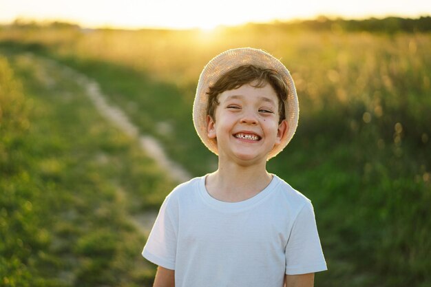 Foto retrato de un niño sonriente con una camiseta blanca y un sombrero jugando al aire libre en el campo al atardecer