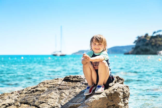 Retrato de un niño sonriente mirando hacia arriba, sentado sobre una roca junto al mar cuando hace calor. Vacaciones familiares de verano en la costa francesa. Niño con camiseta de protección solar y zapatos de natación.