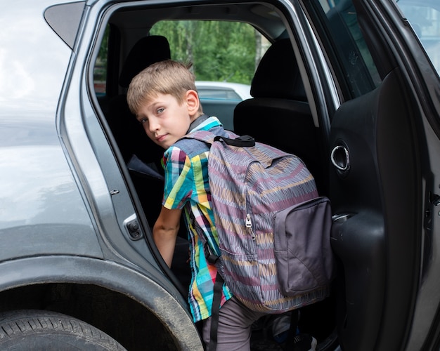 Retrato de niño sonriente con mochila suelta en coche. Llevar a un alumno después de la escuela. El chico se sube al coche.