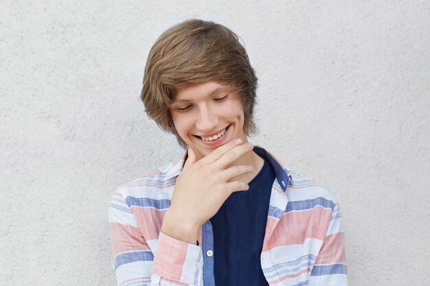 Retrato de niño tímido sonriente con peinado de moda con camisa casual mirando hacia abajo sosteniendo la mano en la barbilla con hoyuelo en la mejilla