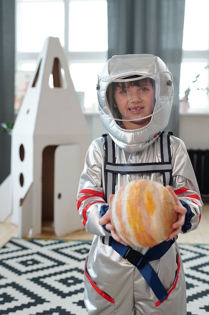 Retrato de niño en traje de astronauta sosteniendo la figura del planeta y sonriendo a la cámara de pie en la habitación