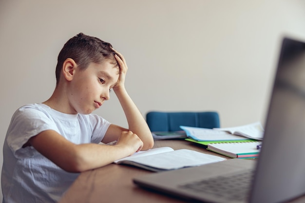 Retrato de un niño triste, cansado y perezoso que tiene una lección de video en línea en una laptop en casa