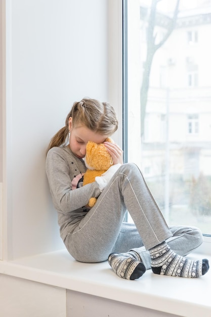 Retrato de un niño triste sentado en el alféizar de la ventana con un juguete suave. La niña está en el hospital con un osito de peluche.