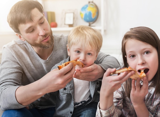 Retrato de niños felices y padre comiendo pizza mientras está sentado en el sofá en casa