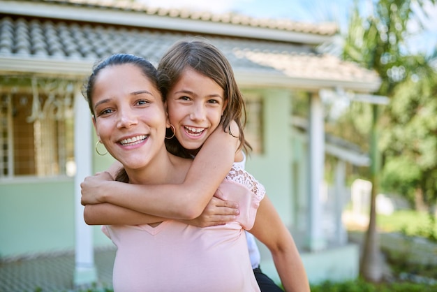 Retrato de niños y una madre a cuestas con su hija al aire libre en el jardín de su casa juntos Cara sonriente y amor con una mujer feliz que lleva a su hija afuera en el patio trasero