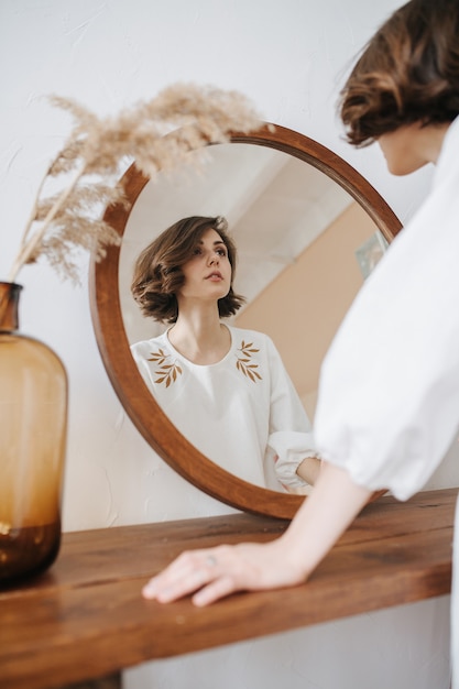 Foto retrato no espelho de uma mulher jovem e bonita e sensual. ela está usando um vestido branco com estampa de folhas. bela morena com cabelo curto dentro de casa. por trás.