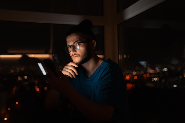 Retrato nocturno de un joven pensativo usando un teléfono inteligente cerca de la ventana