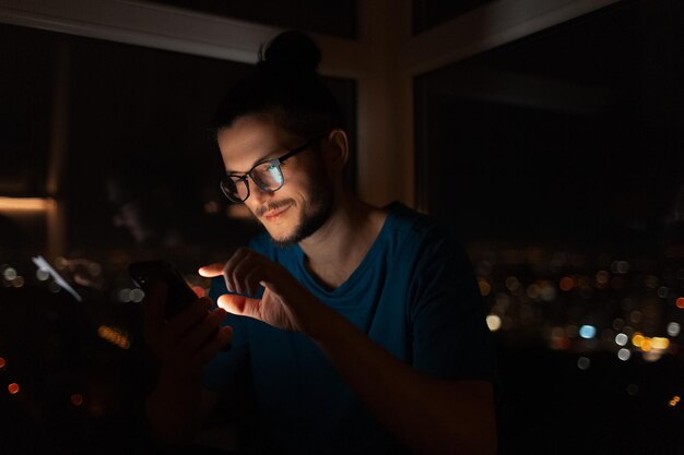 Retrato nocturno de un joven tocando la pantalla del teléfono inteligente Fondo de la ventana