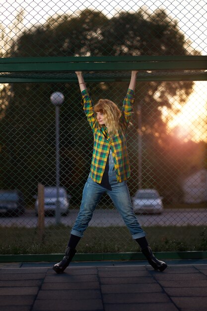 Retrato nocturno de pelirroja jengibre niña sonriente vestida con camisa a cuadros y mono jean, colgando en el campo deportivo al aire libre