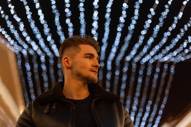 Retrato nocturno urbano de un apuesto hombre hipster con peinado en una chaqueta de cuero negra de moda caminando en la ciudad con luces azules