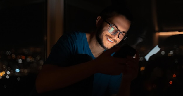 Retrato noturno de homem sorridente usando smartphone perto da janela banner panorâmico