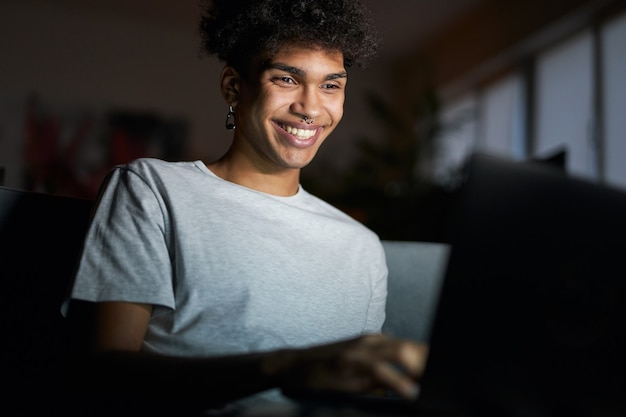 Foto retrato noturno de um jovem sorridente trabalhando em um laptop em um quarto escuro, sentado em um sofá em casa