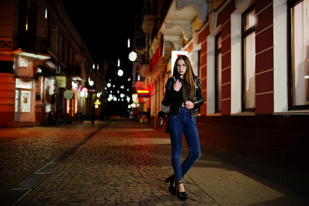 Retrato noturno do modelo de menina vestindo óculos jeans e jaqueta de couro com mochila contra as luzes das ruas da cidade