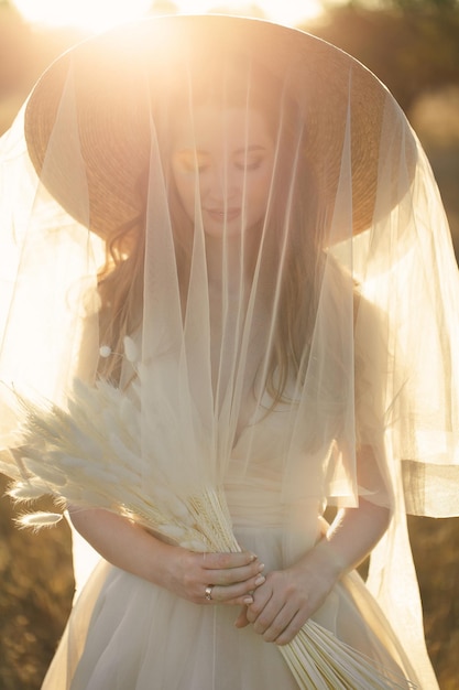 Retrato de una novia gentil con un sombrero de paja y un velo de velo al atardecer