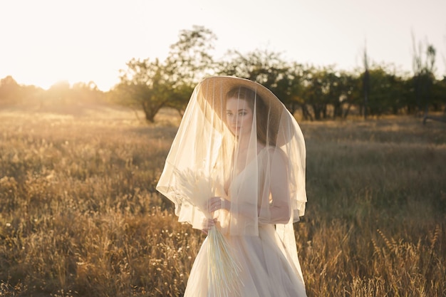 Retrato de una novia gentil con un sombrero de paja y un velo de velo al atardecer