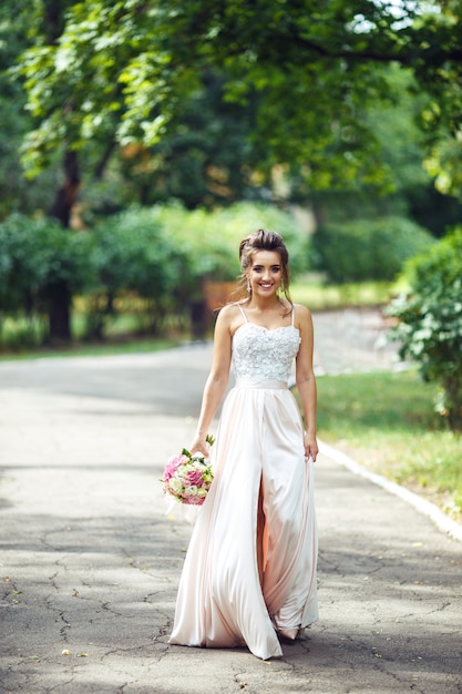 Retrato de novia hermosa con un ramo de novia para un paseo por el parque. La mujer feliz del recién casado se coloca entre arbustos verdes en el jardín. Novia sonriente Día de la boda. Novia de moda.