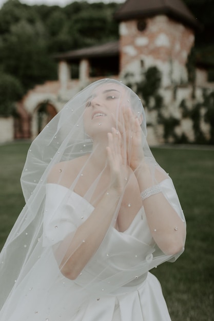 Retrato de novia hermosa en vestido de novia beige con peinado rizado moderno y velo caminando sobre g
