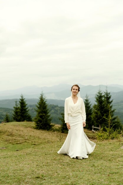 Retrato de novia Una joven con un vestido de novia blanco y un ramo de flores y vegetación en las manos contra el telón de fondo de montañas y bosques al atardecer