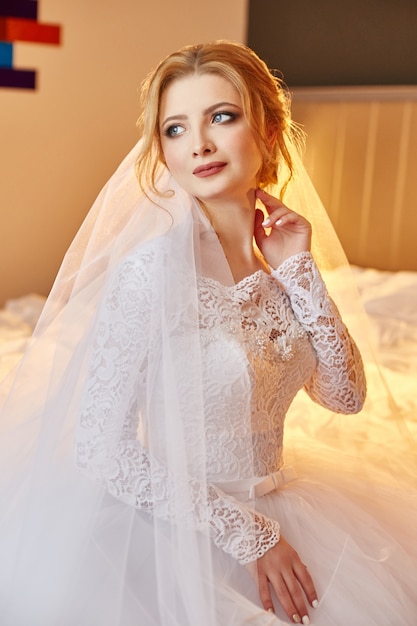 Retrato de una novia sentada en un elegante vestido de novia blanco en la cama y preparándose para la ceremonia de la boda. mujer rubia con un vestido blanco y un velo en la cabeza. La novia está esperando a su novio