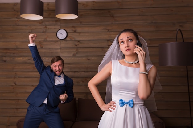 Retrato de novia con teléfono móvil contra el novio feliz en la habitación de madera