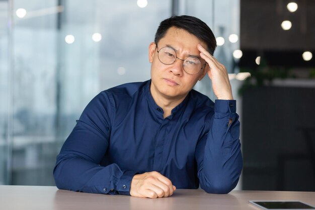 Retrato de oficinista con exceso de trabajo y cansado en el lugar de trabajo hombre asiático con gafas y camisa mirando