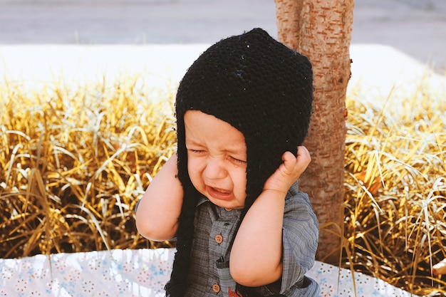 Retrato de otoño de 2-3 años de edad niño llorando en el jardín. Temporada de otoño. Infeliz bebé de raza mixta con gorro de punto