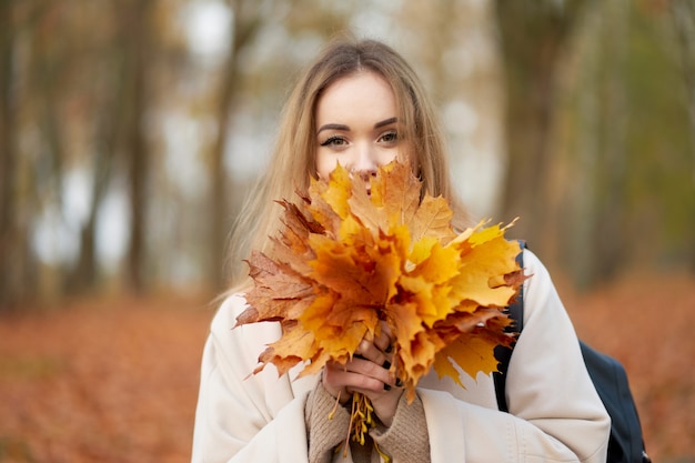 Retrato de otoño. Hermosa chica rubia con estilo con ramo de hojas de arce cerca de la cara con abrigo de otoño de moda posando en el parque de otoño.