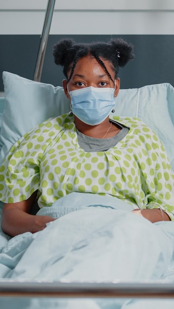 Retrato de un paciente afroamericano con mascarilla y esperando recibir asistencia médica y tratamiento contra la enfermedad. Mujer enferma sentada en la sala del hospital durante la pandemia.