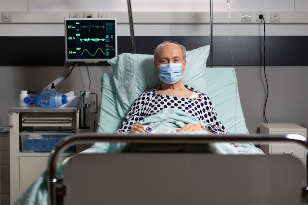 Foto retrato de paciente anciano enfermo con máscara quirúrgica descansando en la cama de un hospital