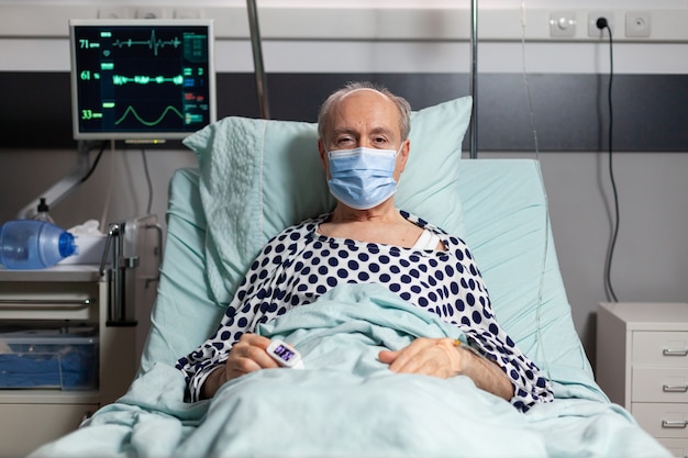 Foto retrato de paciente anciano enfermo con máscara quirúrgica descansando en la cama de un hospital