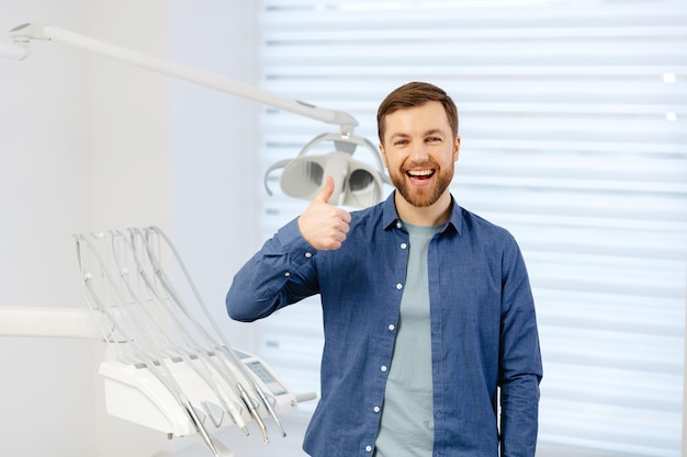 Retrato de paciente feliz cerca de la silla dental y muestra un gesto con un pulgar hacia arriba clase buena Odontología moderna con el uso de nuevas tecnologías