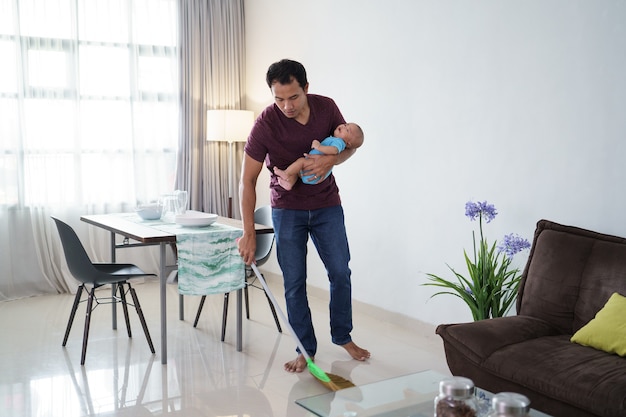 Retrato de padre asiático limpiando el piso con escoba mientras lleva a su bebé