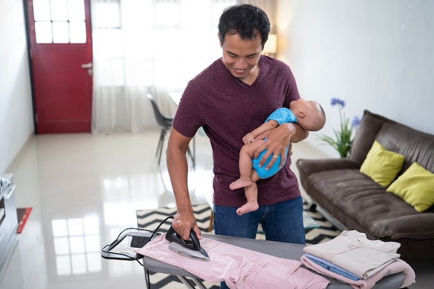 Retrato de padre asiático planchando su ropa mientras sostiene a su bebé en su mano