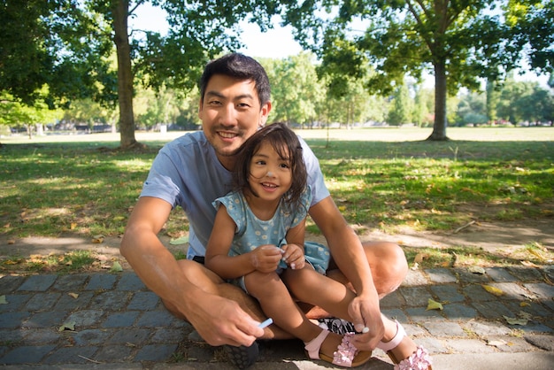 Retrato de un padre asiático posando con su hija para la cámara. Hombre sonriente sosteniendo crayones de colores y una niña sentada en el pavimento en el parque de la ciudad mirando la cámara. Descanso de verano, concepto de paternidad.