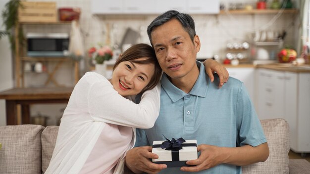 retrato de padre jubilado asiático e hija adulta mirando la cámara con el regalo del Día del Padre en la sala de estar en casa. la mujer sonriente abraza al hombre y se apoya en su hombro