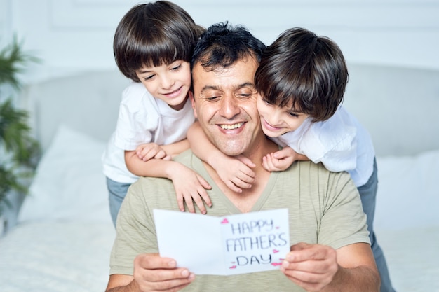 Retrato de padre latino que parece feliz mientras sus dos niños pequeños abrazan a su papá, dándole una postal hecha a mano, saludando con el día del padre, pasando tiempo juntos en casa. Paternidad, hijos