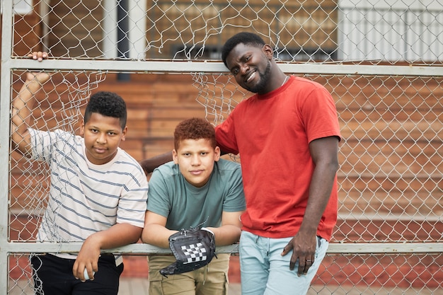 Retrato de padre negro positivo posando con hijos adolescentes con guante de béisbol y pelota, valla de red metálica con orificio