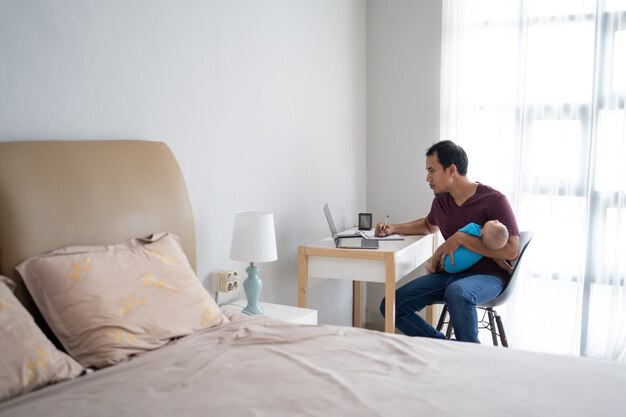 Retrato de un padre ocupado trabajando desde casa mientras sostiene a su bebé bebé