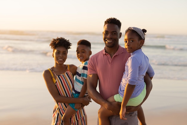 Retrato de padres jóvenes afroamericanos sonrientes que llevan a su hijo e hija contra el mar y el cielo