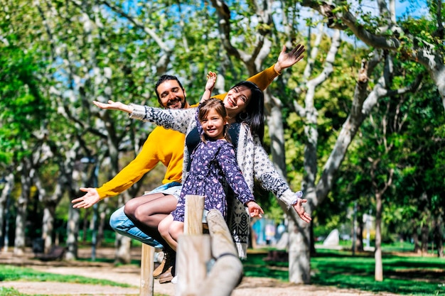 Retrato de padres sonrientes e hija con los brazos extendidos sentados en la barandilla contra los árboles en el parque