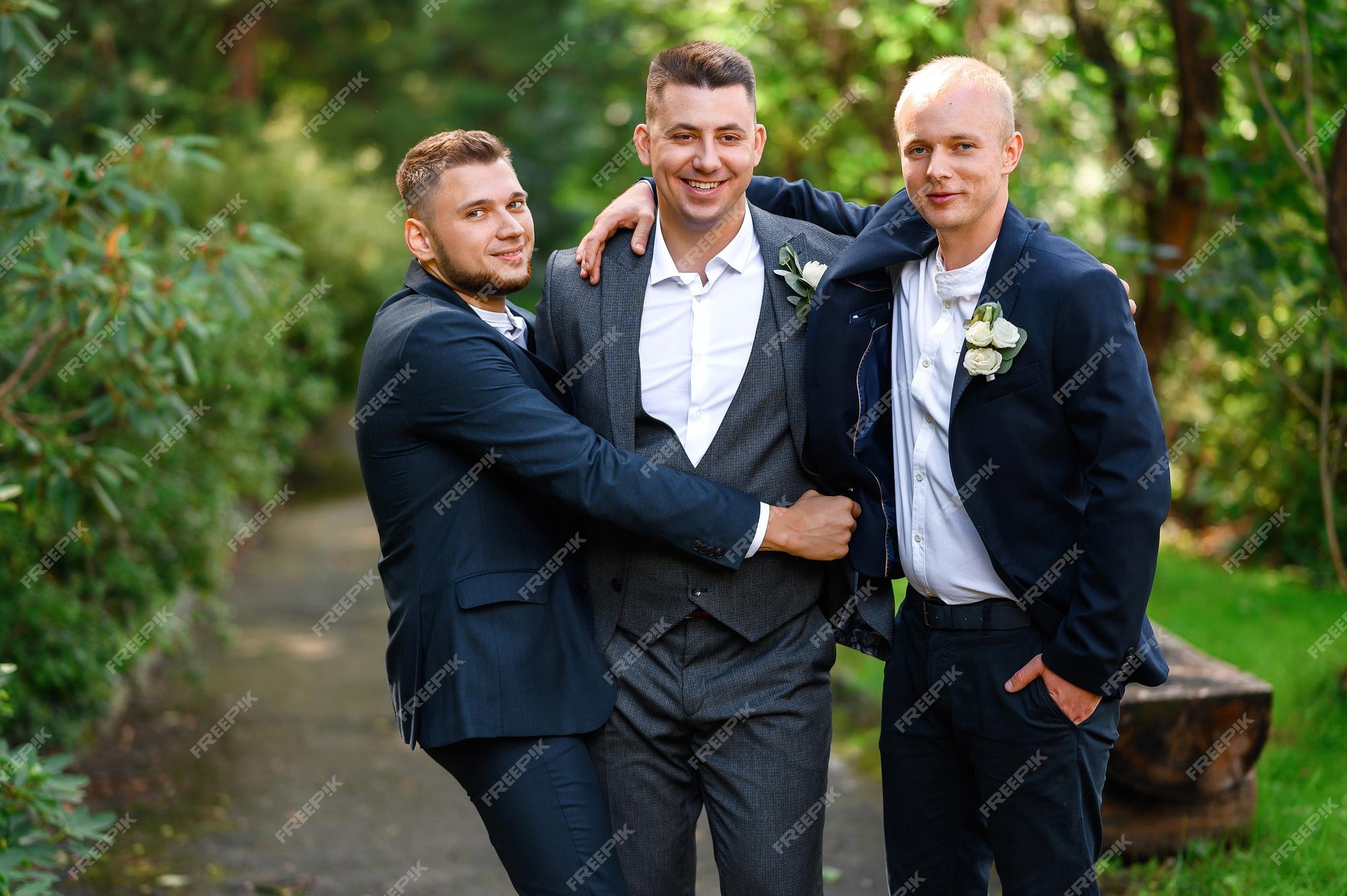 Retrato de padrinos de boda atractivos en traje azul oscuro abrazando al novio a la cámara mientras posa en el jardín amigos hombres de novio saludando con el día de la