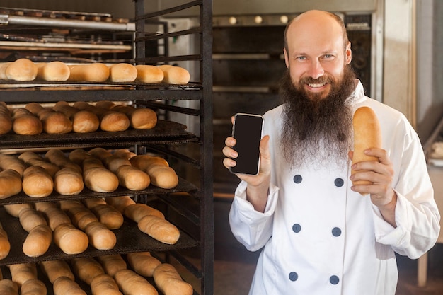 Retrato de panadero adulto satisfecho con barba larga en uniforme blanco de pie en la panadería y sosteniendo el teléfono y pan fresco, mirando a la cámara con una sonrisa con dientes, Interior, concepto de profesión